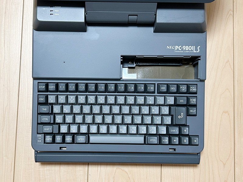 キーボード部分を真上から見た状態です。かなりコンパクトになっているものの、基本的なキーに関しては問題無くタイピングすることができました。キーボード右上部分には、メモリを増設するためのスロットが用意されています。蓋を閉めると左側に電源とFDDのインジケータが見えますが、これは稼働中にも見えるようになっていて、FDDへアクセスしていることは右側面のFDDを見なくてもわかるようになっていました。手前に引き出されているのが取っ手で、かなりしっかりしています。