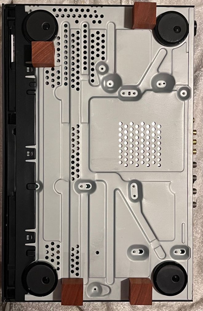 BDレコーダーの底面に「木のサイコロ」(支持脚)を使う場合。付属の「偏心インシュレーター」は背が低いので、30mm角程度でも浮かせることができる。ただし底面は凸凹していて同じ高さの面で元の脚部に近い位置を選ぶのが難しい。また、底板の直近に回路基板がある箇所があり、凹むと危険だ。この例では本体重量6kg程度のBDレコーダーなのでなんとかなりそうだが、本来は元の脚部を外して置き換える方が安全だ