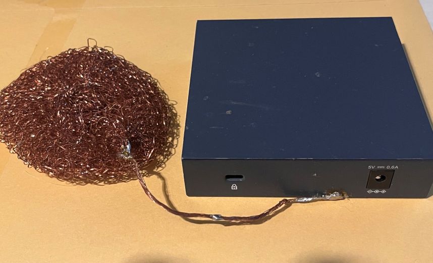 スイッチングハブの背面にはDC5VのDC入力端子がある。これを使って各種電源を試せる。DC5Vの充電池パックを使うと、画質は最高となり、これを基準にして付属のスイッチング電源式ACアダプターのノイズ対策の効果を確認する。ただし音質はクリアーになるもののもうひとつ。左の銅たわしはハブの筐体に接続してノイズを逃がす目的。つまり「床アース」だ。ただしはんだ付けすると改造になってしまうことに留意されたい。ねじ止めを利用するのが無難だ