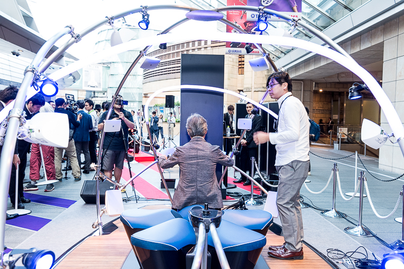 スピーカーをドーム状に配置した空間に座って操作する。手前の黒い突起部分は電動車いすの操縦スティックとなっており、音量を調節できる