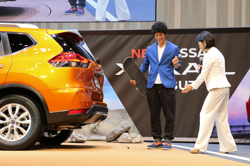 リモコンオートバックドアについて説明され、車両のインテリジェントキーが星野専務の手で渡部さんのポケットに入れられる。右手にはスケートボード、左手にマイクと、バックドアを操作できない状態