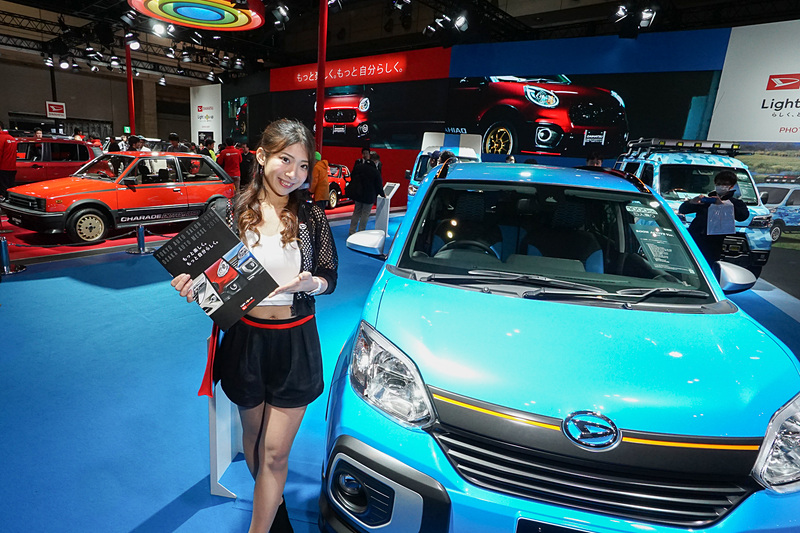 ダイハツブース。「ブーン」ベースでスポーツを楽しむスタイリッシュ系をイメージした参考出品車「BOON ACTIVE Ver.（ブーン アクティブ バージョン）」の前で。奥に往年のホットハッチ「シャレード デ・トマソ ターボ」が見える