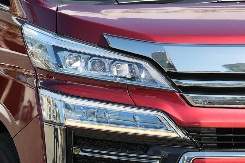 ヴェルファイアではフロントグリルをボディ同色部分で上下に分割し、2段式のヘッドライトデザインを強調。ヘッドライトはアルファードと同じく3眼LEDヘッドライト＋LEDクリアランスランプを標準装備