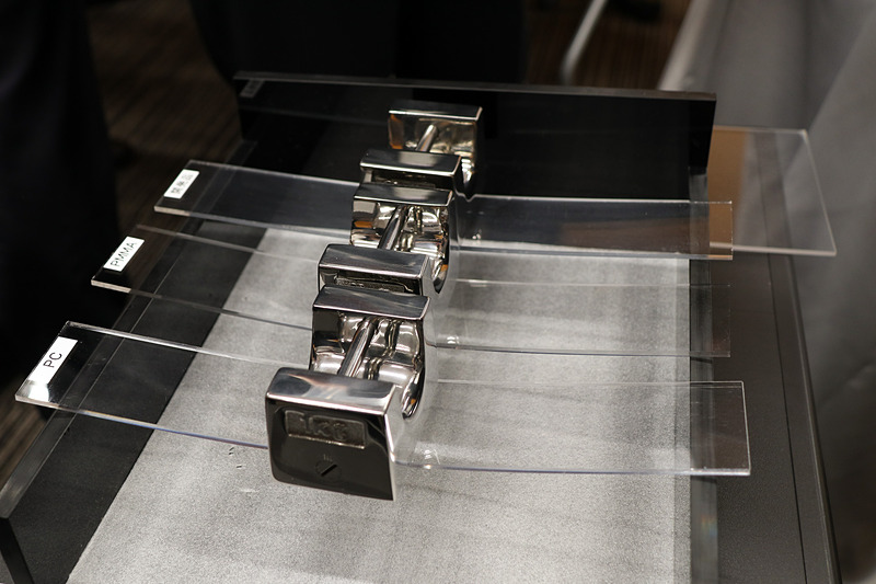 住友化学は2種類のデモを実施。部材のたわみ度合いの比較では、ポリカーボネート、従来型PMMA、PMMAベースの透明樹脂の3種類に1kgのウエイトを乗せて沈み込み具合を表現