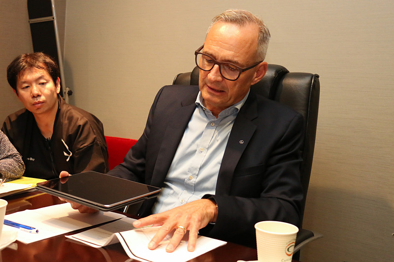 手元にあった紙資料を曲げた模擬の路面バンプとタブレットを使い、マジックボディコントロールとDYNAMIC BODY CONTROLサスペンションの違いを解説するフリュー氏