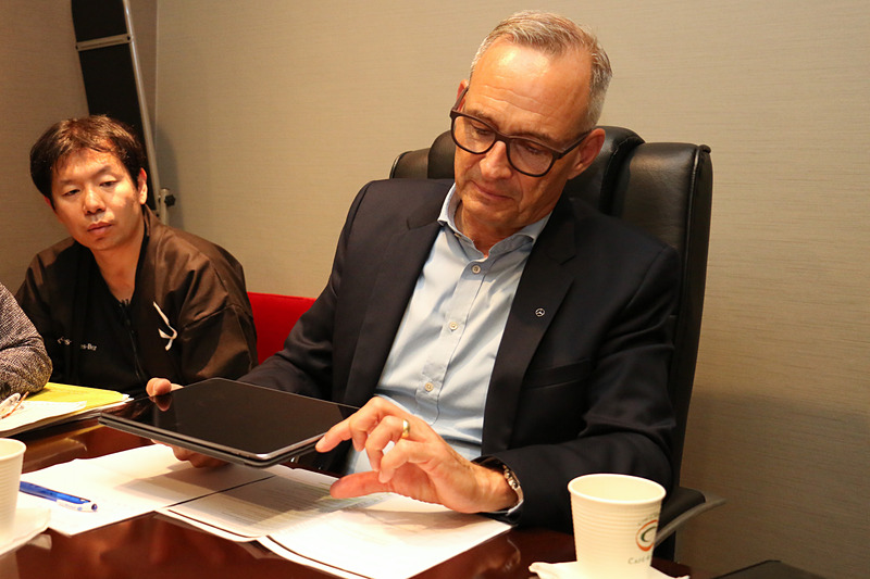 手元にあった紙資料を曲げた模擬の路面バンプとタブレットを使い、マジックボディコントロールとDYNAMIC BODY CONTROLサスペンションの違いを解説するフリュー氏