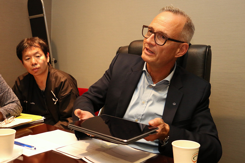 手元にあった紙資料を曲げた模擬の路面バンプとタブレットを使い、マジックボディコントロールとDYNAMIC BODY CONTROLサスペンションの違いを解説するフリュー氏