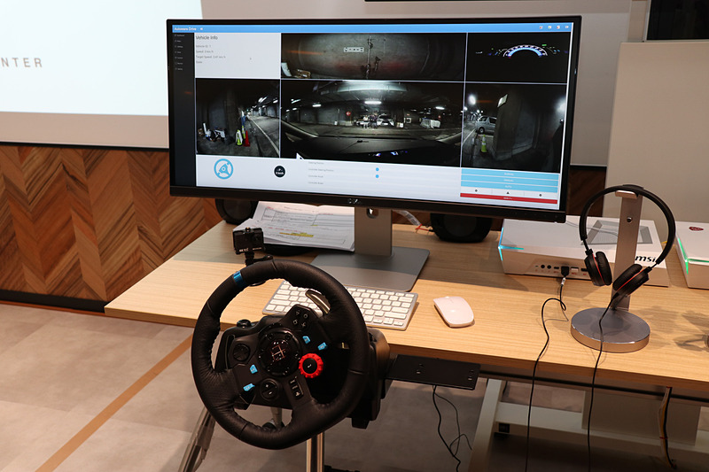 遠隔運転担当者の席にはPCとワイドディスプレイに加え、ゲーム用のステアリングコントローラーを設置。ディスプレイの表示はクルマの運転席を模して、中央に大きく車両正面を表示するほか、上側に車両後方、両サイドにドアミラーのように車両側面、右上にメーターパネルが映し出されている