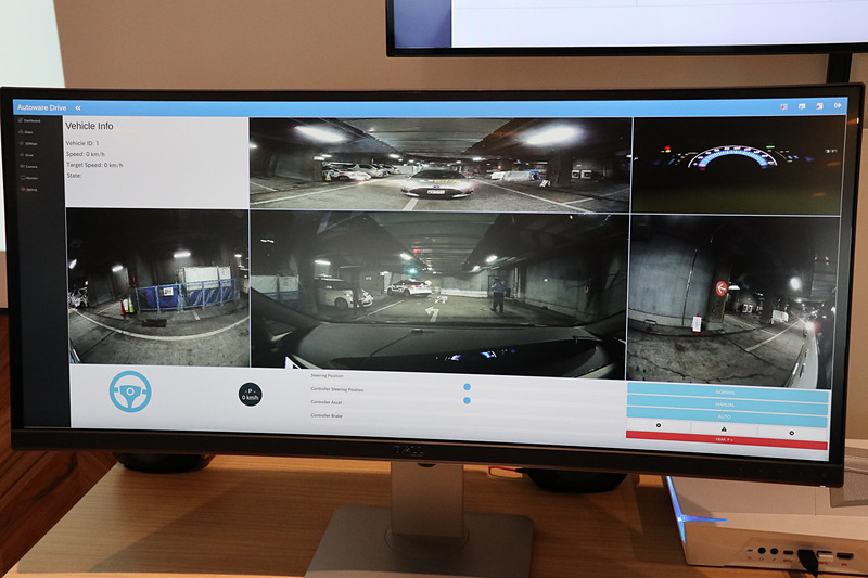 遠隔運転担当者の席にはPCとワイドディスプレイに加え、ゲーム用のステアリングコントローラーを設置。ディスプレイの表示はクルマの運転席を模して、中央に大きく車両正面を表示するほか、上側に車両後方、両サイドにドアミラーのように車両側面、右上にメーターパネルが映し出されている