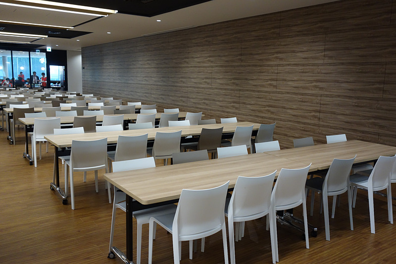 2階の食堂では食事する人数に合わせてさまざまな形状のテーブルが用意される。モニターにはメニューとともに残数の表示も。ユニークなのはフロアに書かれた英数文字。この日はAメニューが「カレー」（300円）だったが、「A/56」と書かれたところは「カレーに56人が並んでいます」という意味。ひと目で何のメニューにどれくらいの人が並んでいるかが分かるようになっている