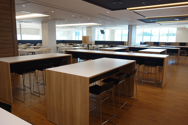 2階の食堂では食事する人数に合わせてさまざまな形状のテーブルが用意される。モニターにはメニューとともに残数の表示も。ユニークなのはフロアに書かれた英数文字。この日はAメニューが「カレー」（300円）だったが、「A/56」と書かれたところは「カレーに56人が並んでいます」という意味。ひと目で何のメニューにどれくらいの人が並んでいるかが分かるようになっている