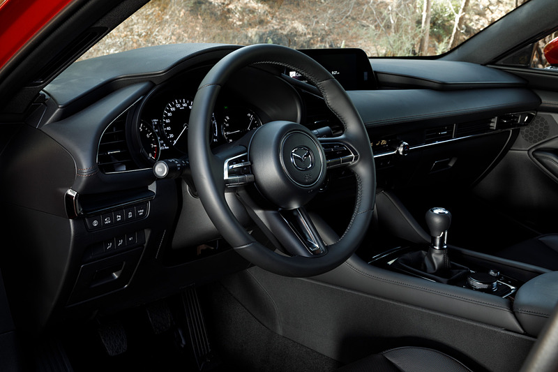 5ドアハッチバックのインテリア。新型Mazda3のコクピットはすべてをドライバーを中心に左右対称にレイアウト。ステアリングスポークやセンターディスプレイなど、パーツ単位で「バニシングポイント」（遠近法で言う収束点）に基づいてデザイン。また、本革にはマツダ独自のシボを採用したほか、シフトパネルにはレーザー加工で柄を刻み込んだ黒のメタリック層と、まっすぐ光を透過・減衰させるカラークリア層からなる2層成形と呼ばれる技術を採用