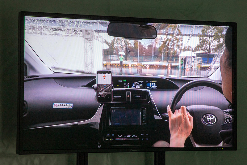 クルマと歩行者の両方に自らの位置を発信する装置を付けて、お互いが通信しあうことで、物陰でも接近が感知できるシステムのデモ。歩行者として体験できる