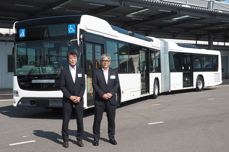開発した「ハイブリッド連節バス」の前に立つ、日野自動車株式会社 車両企画部 チーフエンジニア 山口誠一氏（左）と、いすゞ自動車株式会社 バス商品企画・設計部チーフエンジニア 鈴木隆史氏（右）