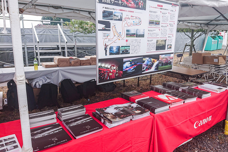 貸し出しブースのテーブルにはEOS 90Dのカタログや写真展のチラシなどが並べられていた。吊されたパネルは熱田氏の連載が掲載されているデジタルカメラマガジンの2018年12月号に掲載されたF1特集ページをパネル化したもの