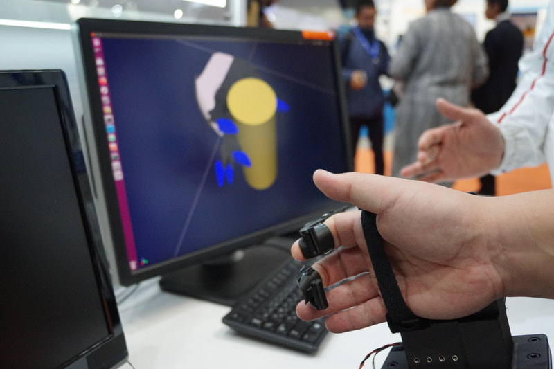 トヨタブースに展示された力覚提示可能なロボットハンド操縦装置。モニターに表示されているゴムの棒を握る感覚をロボットハンドで再現
