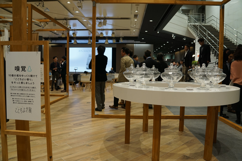 嗅覚ブース「ここちよい香り」。選択肢は「フルーツとライム」「もみの木とお香」など10種類（監修：生活の木）。円形のテーブルに並べられた10本の瓶。フタを開けると、柑橘・ひのき・ハーブなど、さまざまな香りがふわりと漂う。この中からここちよい香りを選択する