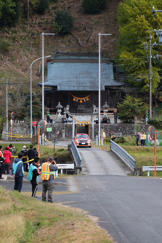 神社を背景に走るラリーカー。黄色、青、緑のタバードを付けているのは報道陣。橋の手前左側や神社の境内にいる人は観戦客や地元住民