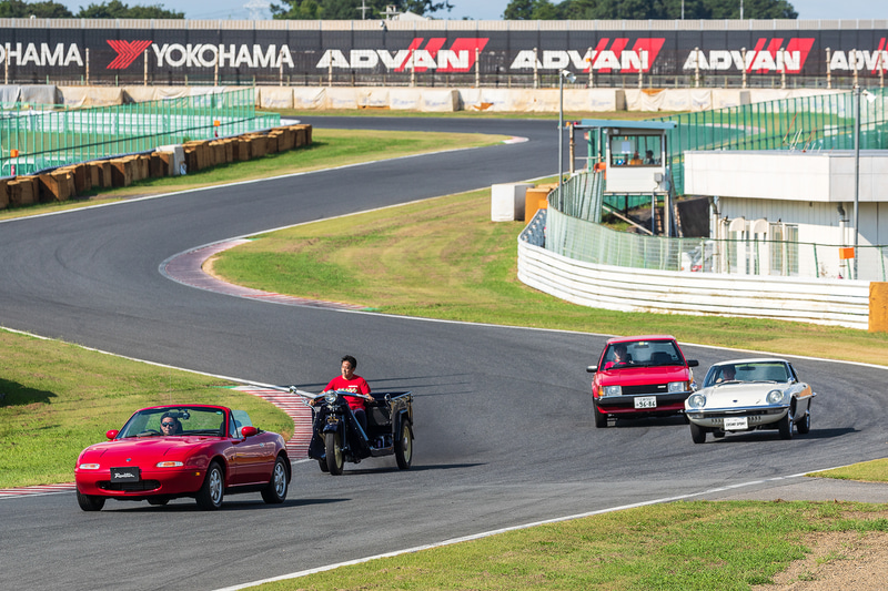 歴代のクルマのパレード。先頭は1989年発売の初代「ロードスター」、順に、1947年式の三輪トラック「GA型（通称グリーンパネル）」、1967年発売の「コスモスポーツ」、1980年発売の初代FF「ファミリア」