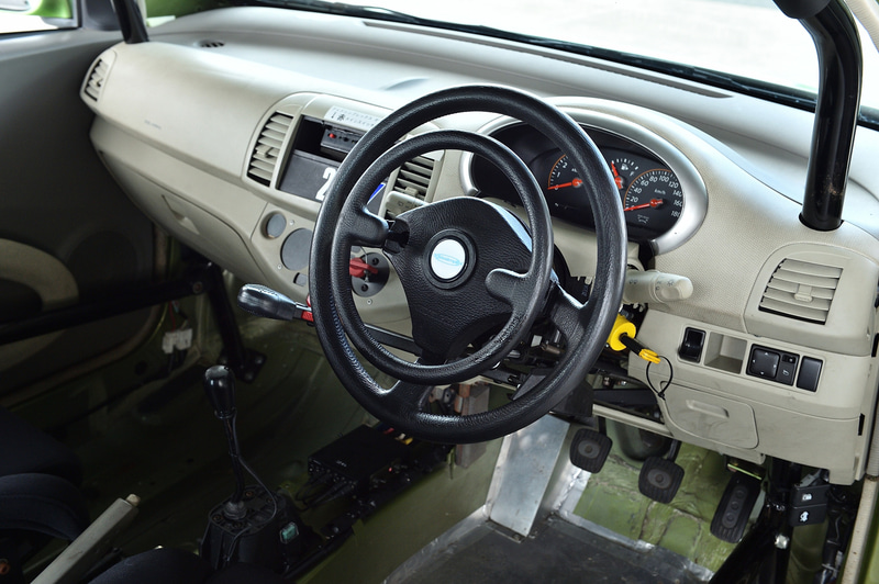 グイドシンプレックス製の手動運転装置。ハンドル左のコラムシフトのようなレバーを前に押すことでブレーキ、ハンドル中央のリングを押し込むことでアクセル