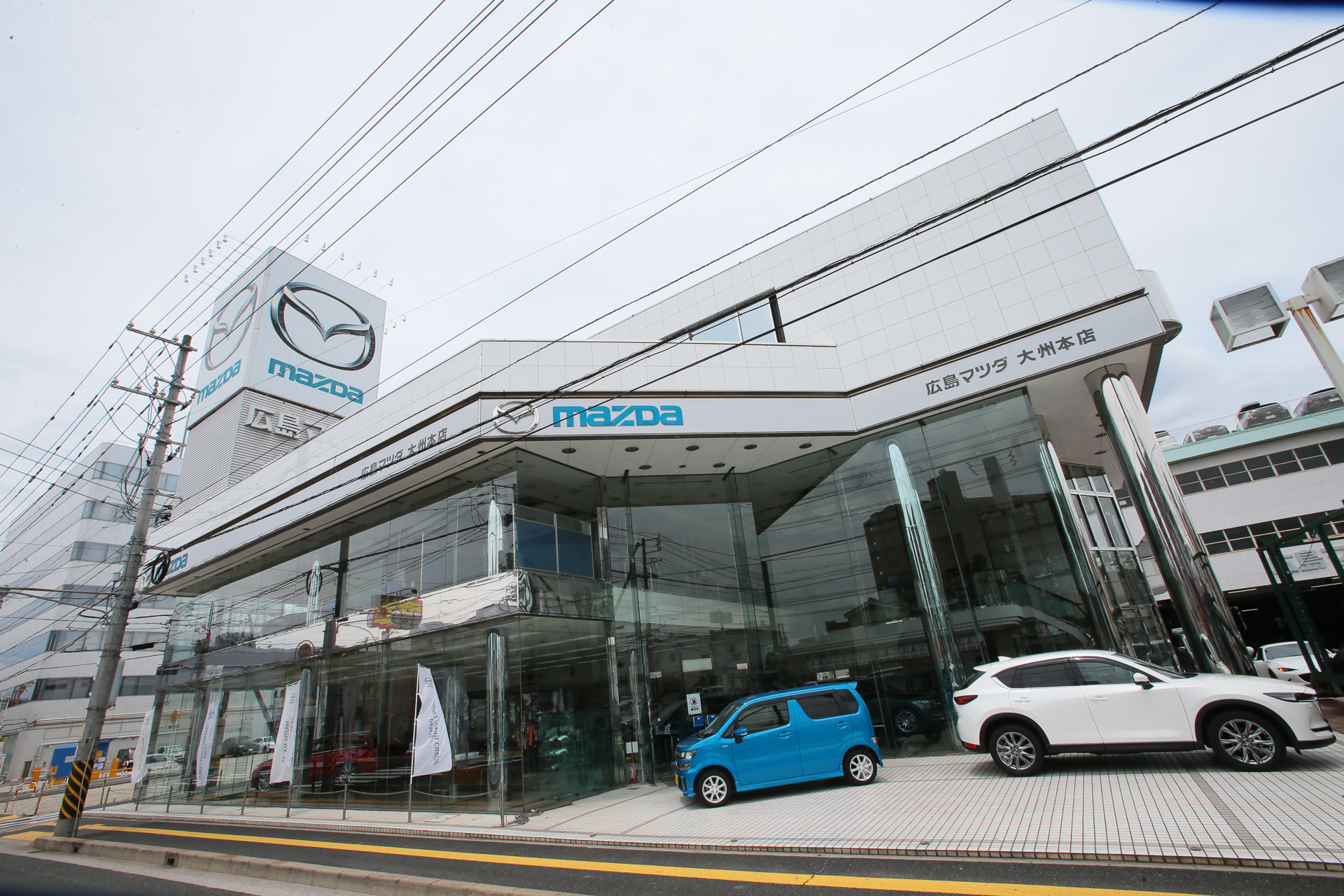 HM RACERSの活動拠点になる広島マツダ 大州本店（広島県広島市南区大州4-10-11）。マツダ車の販売、アフターサービスをはじめ、2階にはHM RACERSの活動拠点を構え、オリジナルブランドのチューニングパーツやアフターパーツの販売を行なっている