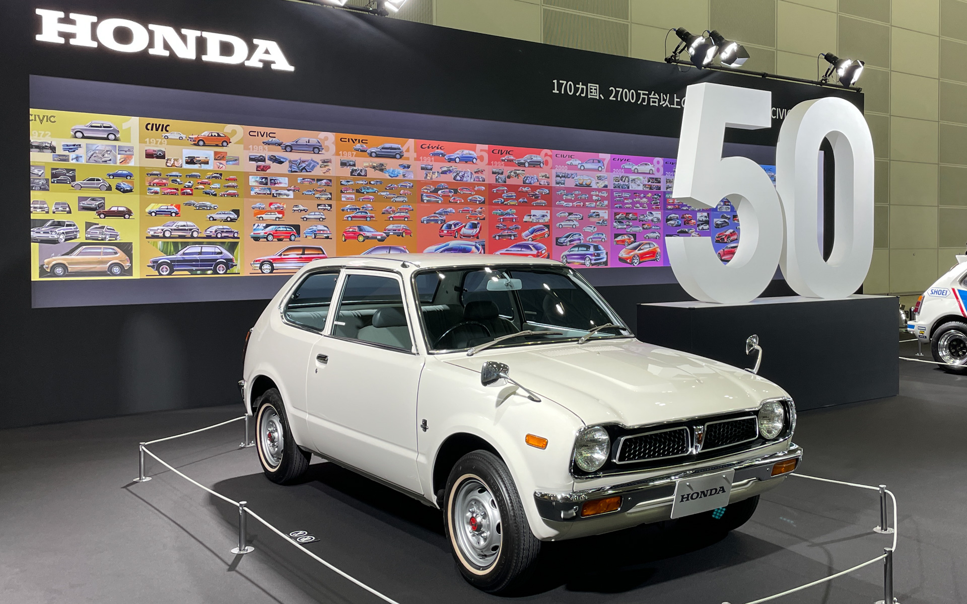 シビック50周年の歴史を伝えるホンダブース。1972 初代 CIVIC（2ドア）のうしろには歴代CIVIC デザインスケッチギャラリーを展示。歴代シビックのデザインスケッチをそれぞれの世代ごとにまとめた11枚の連作が公開された