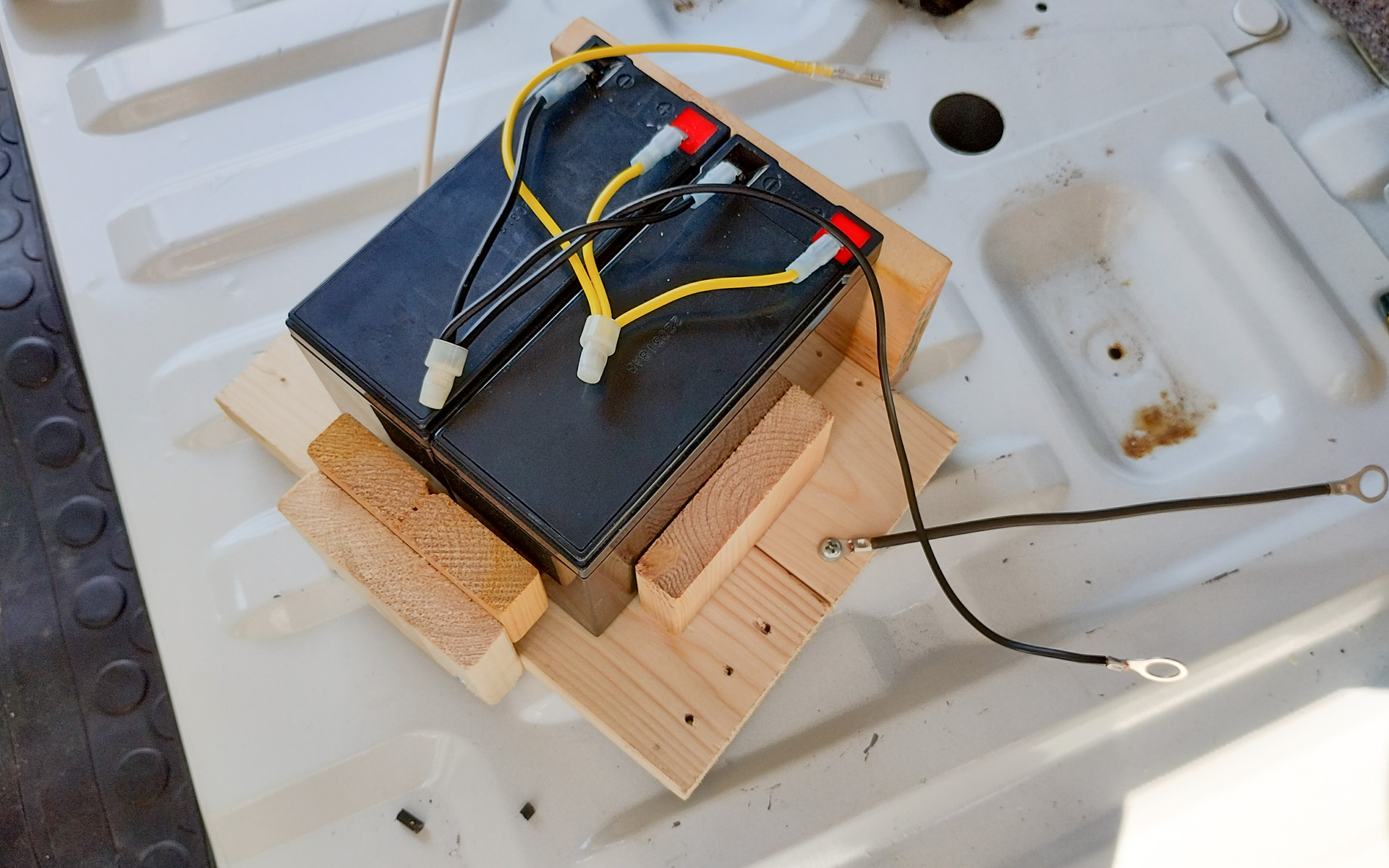 用意したバッテリと木で作った台座。これでバッテリは倒れない。シート下パネルを固定するボルトにワイヤー（電線）に共締めして台座を固定する
