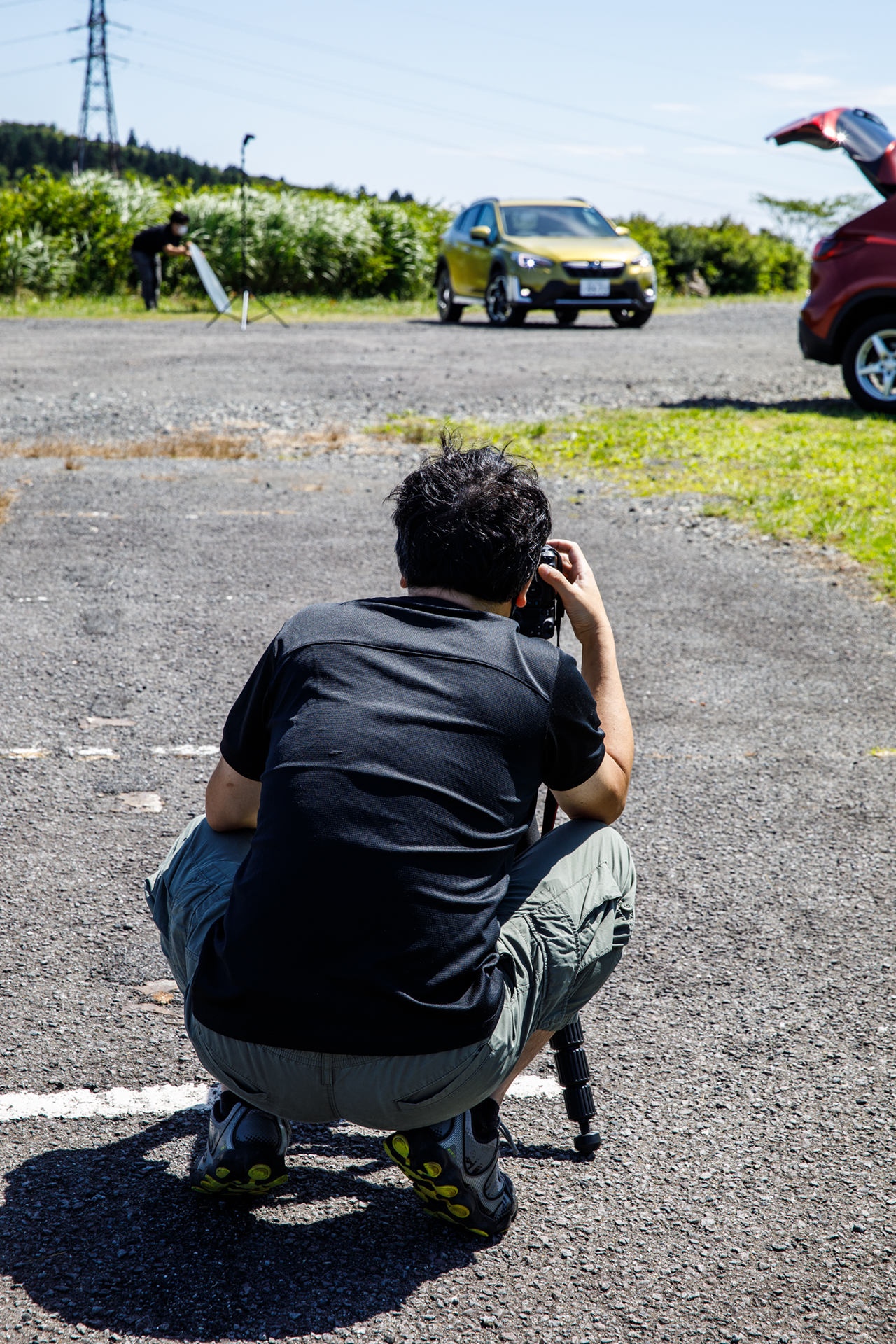 逆光の条件で撮影する安田カメラマン。レフ板だけでは自動車の後部しか明るくできないので、ワイヤレスでコントロールできるフラッシュも使っている。アシスタントが多ければ複数のレフ板を使うこともあるそうだ。望遠レンズでの撮影なので、やはり自動車から離れて撮影する
