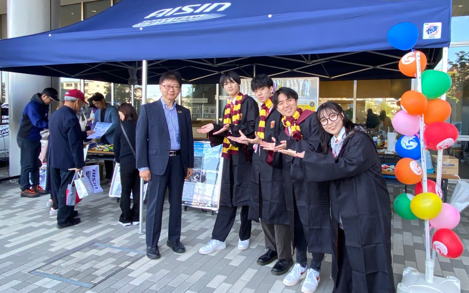 ずっと頑張ってきた共同研究プロジェクトのブースの前で、ハリーポッターの衣装で揃えた立命館大学経営学部・徳田昭雄ゼミ生のみなさん。右から田中さん、木谷さん、林さん、佐藤さんは、応援に駆けつけてくれたアイシンの鈴木研司取締役とも、気さくにいろんな話をしていました。若いって素晴らしい！