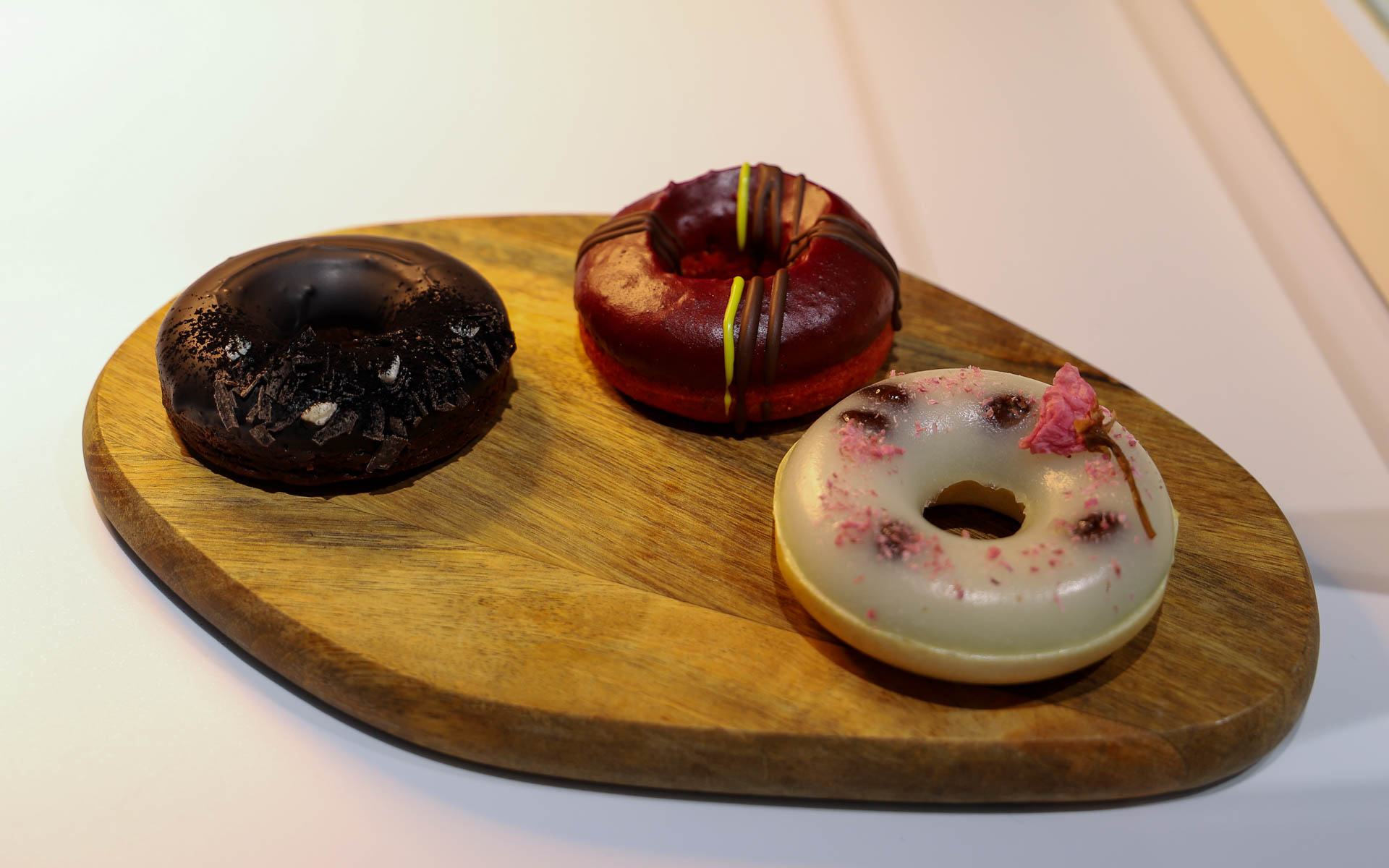 「HOCUS POCUS」による、ニューヨーク、ロンドン、東京の都市をコンセプトにした3種類のオリジナルドーナツ。ニューヨーク（左）は、“ビッグアップル”をイメージした大ぶりのリンゴが入ったチョコレートドーナツ。ロンドン（中央）は、ビーツパウダーで色づけしたレモンケーキのような仕上がりで、タータンチェックをカシスソースでデコレーションした赤いドーナツ。東京（右）は、小豆を入れたお餅の生地に、モナカを採用し、桜の塩漬けのパウダーをかけたドーナツ