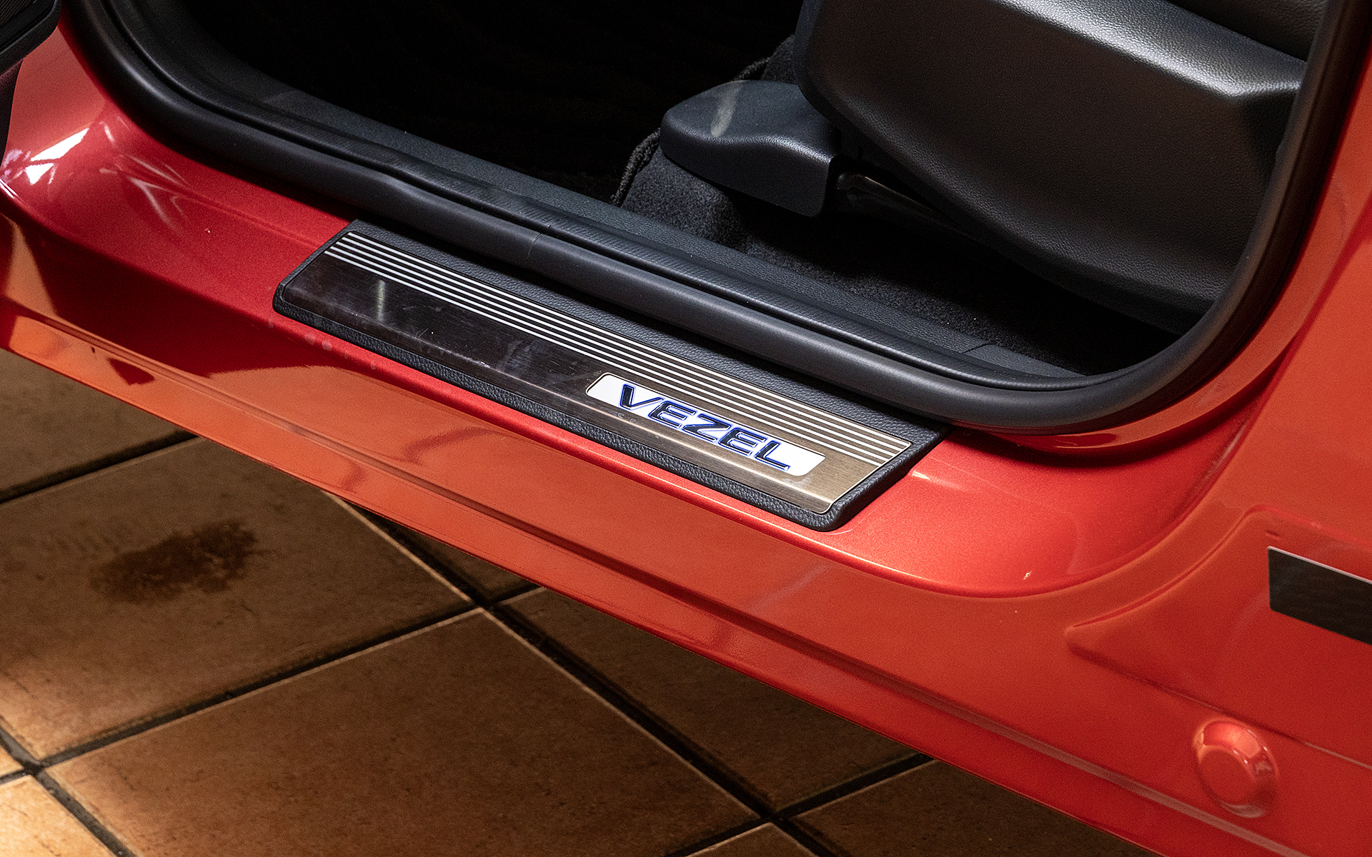 サイドステップガーニッシュ（価格2万9700円）。フロント＆リアの4枚セットで、フロント部はドア開閉連動で光るブルーのLEDイルミネーション付き。材質はステンレスでVEZELロゴ入り。ノーマルは何も付いていないのでステップの塗装保護にもなる