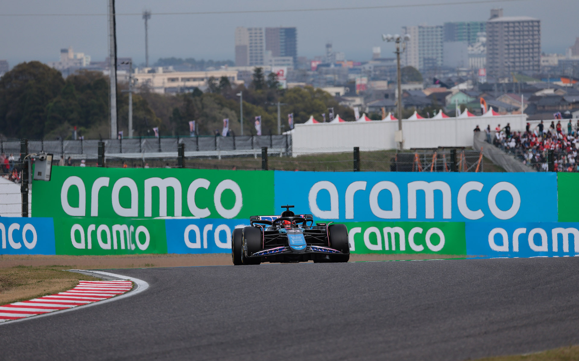 背景はポストの向かって右側の看板と鈴鹿の市街。この看板の後ろに桜の木が並ぶと、逆バンクに進入するマシンの背景に桜を写すことができそう