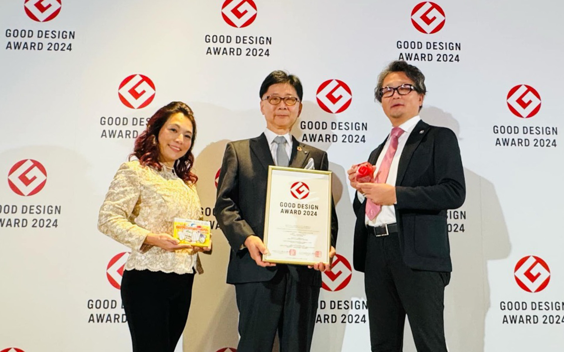 アイシン×立命館大学デザイン科学研究所の共同研究プロジェクトから生まれた「性被害やっつけたるわファミかるた」がグッドデザイン賞を受賞！　おなじみ「Gマーク」のパネルの前で、企画者でありプロジェクトをまとめてきた立命館大学大学院経営学研究科の山田貴子さん（左）と、アイシンの鈴木研司シニアエグゼクティブアドバイザー（中央）、立命館大学副学長の徳田昭雄教授（右）がパチリ