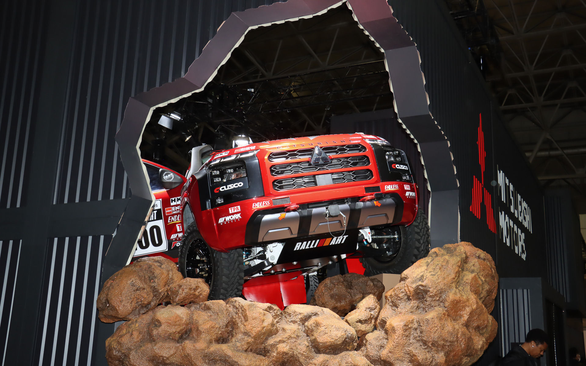 まるでトライトン AXCR 2024 ラリーカーが壁を突き破って飛び出そうとしているような斬新なスタイルで車両展示