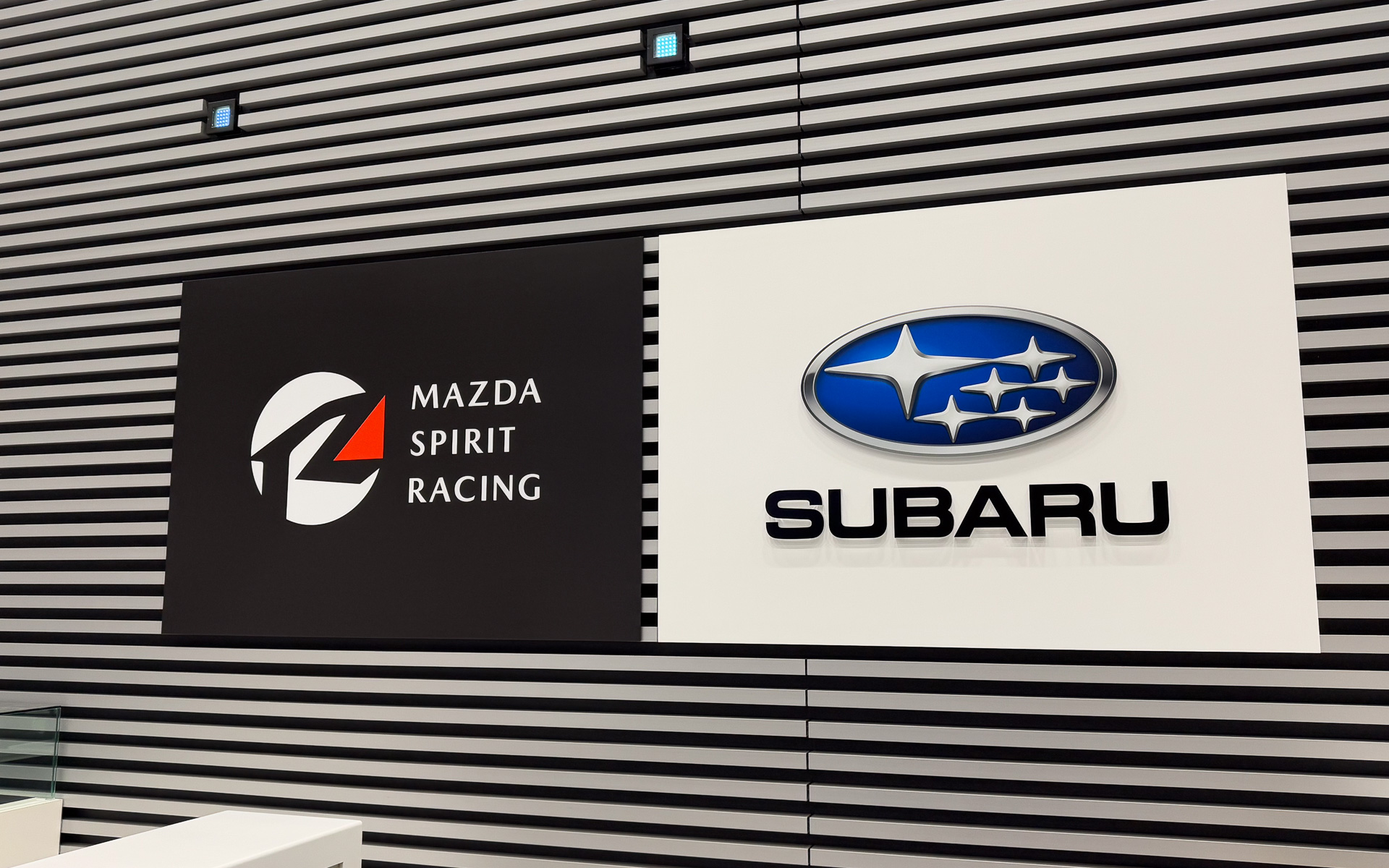 ショールームの受付に掲げられたスバルとマツダ（MAZDA SPIRIT RACING）のロゴ。フォトスポットになるかもしれない