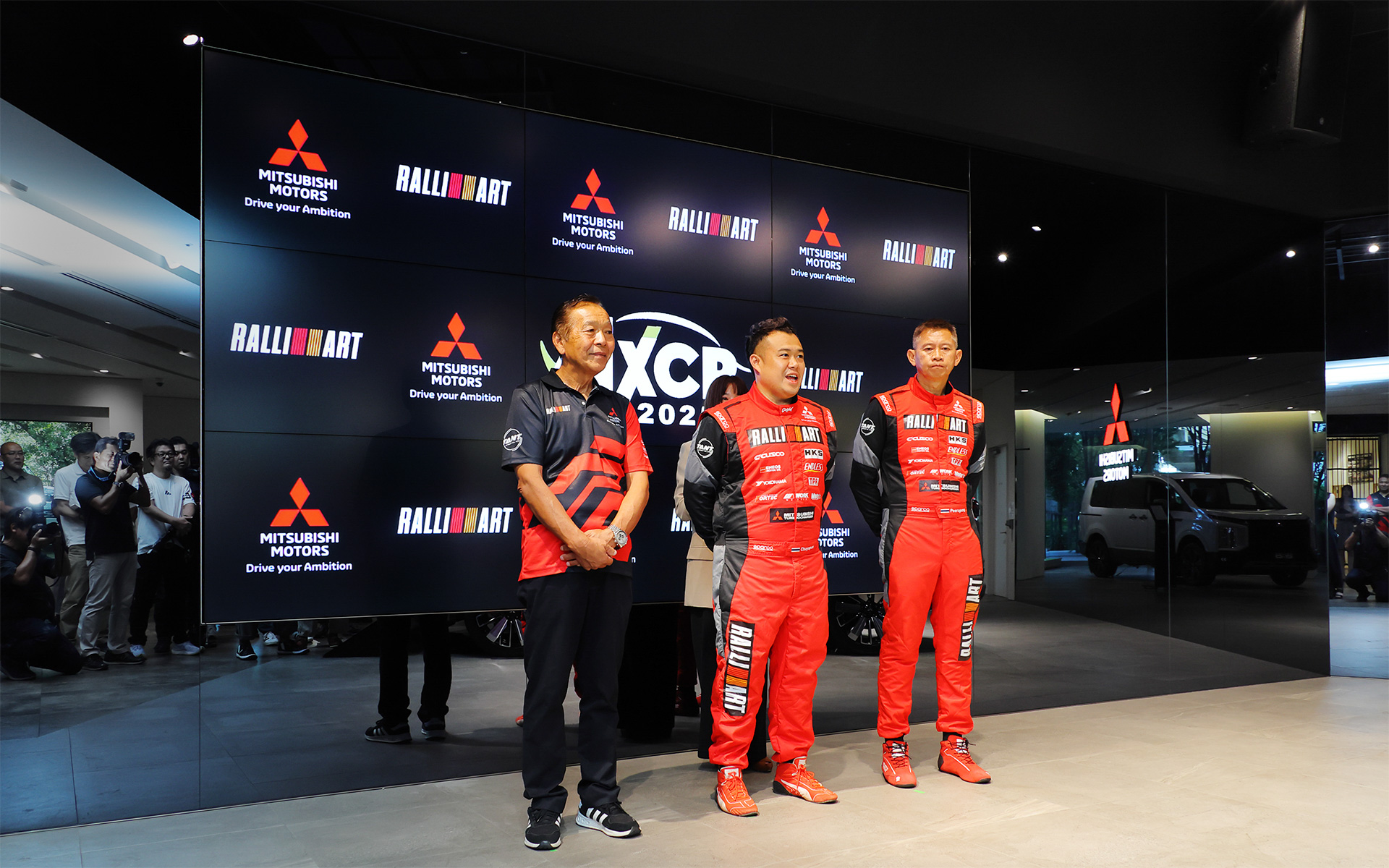 三菱自動車の本社ショールーム（東京都港区）で開催された優勝報告会に登壇した3名。左からチームの総監督を務めた増岡浩氏、ドライバーのチャヤポン・ヨーター選手（タイ）、コ・ドライバーのピーラポン・ソムバットウォン選手（タイ）