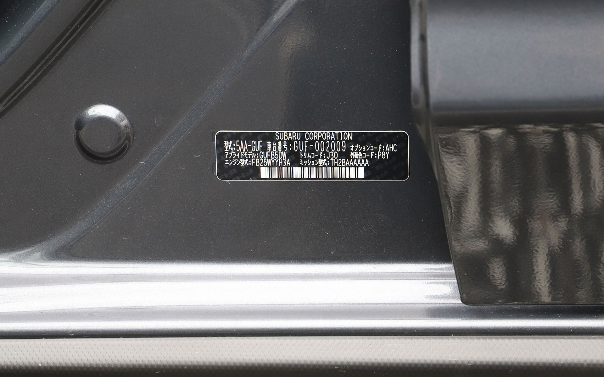 助手席ドアを開けたときに、コーションプレートが車体側に貼り付けられている（今はシールのようなものになっているが、以前は金属の板がエンジンルーム内にあったので、その名残でプレートと呼ばれているのではないかと思われる）。アプライドモデルを左から読み解くと「GU」が2023年から発売されている「インプレッサ」シリーズを表わしており、「F」がハイブリッドであることを示している。次の「B」が発売後に何回モデルチェンジしたかがわかる部分で、Aから始まって1つのモデルではEやFくらいまで変更を重ねていく。「5」はドアの枚数を表わしており、そのあとに続く「DW」は……忘れてしまった。ちなみに、ここに記されているオプションコード（写真だとAHC）をWebサイトで公開されている主要装備表と照らし合わせればどのメーカーオプションが付いているかが確認できるし、外装色コード（写真だとP8Y）はタッチアップペンなどのボディ補修用品を買うときに参考にすると便利だ（編集部撮影）