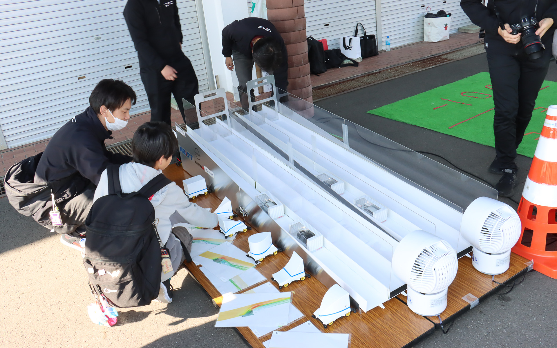 ミニ四駆のシャシーを流用し、重量と前面投影面積を同一条件に合わせ、カウルの形状だけを変更した6種類のマシンを用意。サーキュレーターで強い向かい風を発生させた斜面をどのマシンが一番速く駆け下りるかを予想してもらい、来場者に空気抵抗が車体に与える影響を体感してもらう新たな取り組み