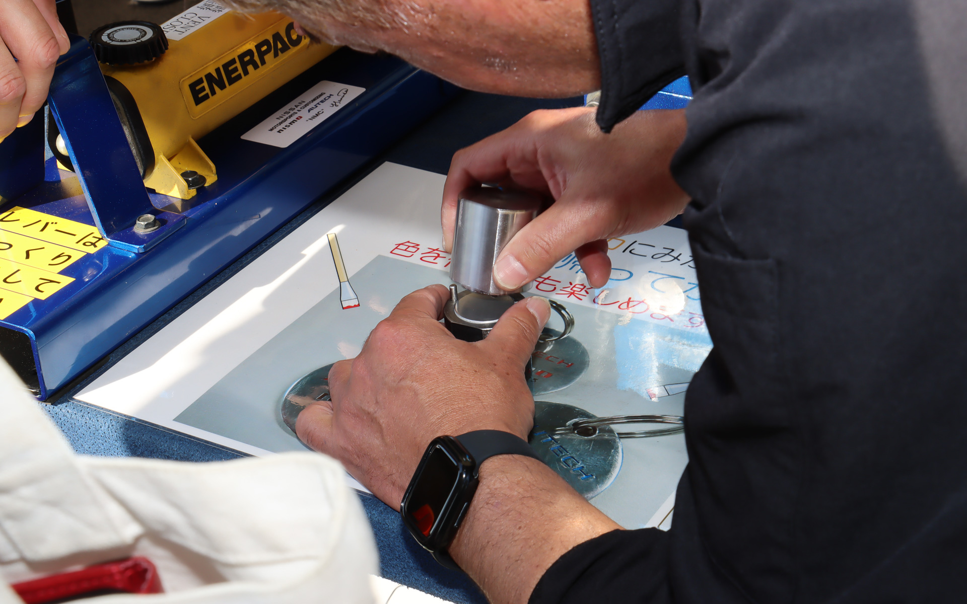 モノづくり体験ブースではイベント参加の記念品にもなるメダル作成を実施。事前に打ち抜かれて片面に刻印したメダルの無地面に油圧プレスでロゴを刻印したあと、紙やすりと研磨剤を使ってピカピカに磨き上げていく