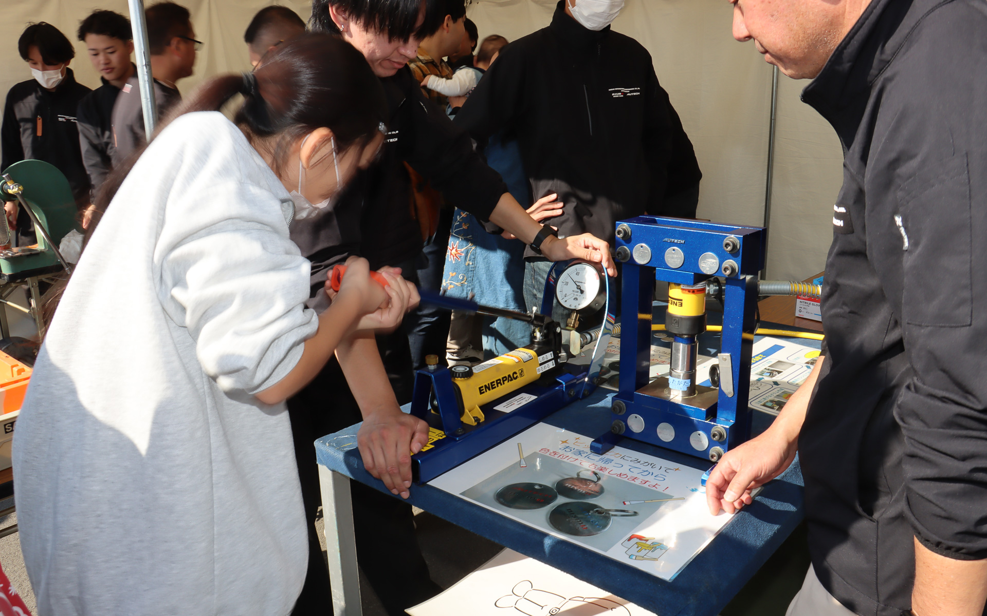 モノづくり体験ブースではイベント参加の記念品にもなるメダル作成を実施。事前に打ち抜かれて片面に刻印したメダルの無地面に油圧プレスでロゴを刻印したあと、紙やすりと研磨剤を使ってピカピカに磨き上げていく