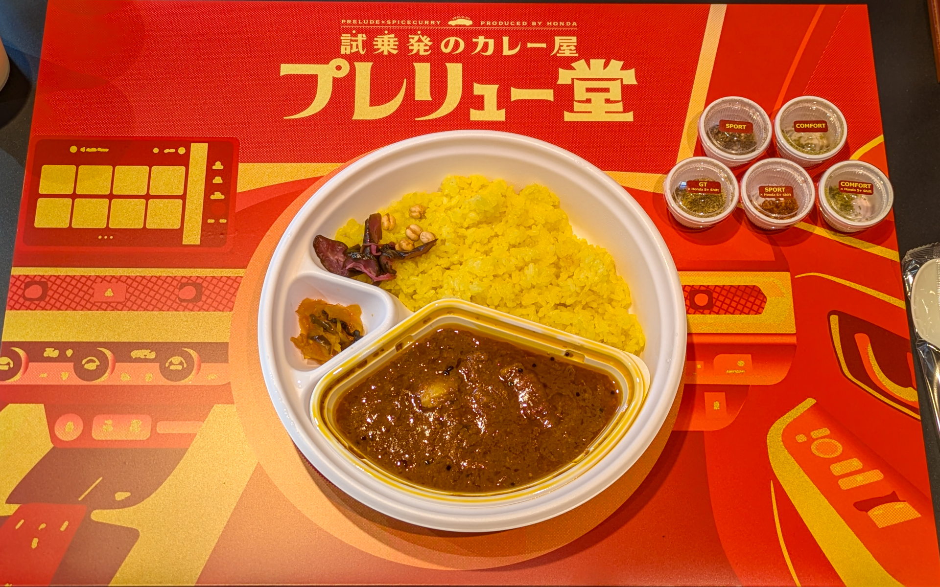 ベースとなるカレー（GT）は、伸びやかで爽やかな走りをトマトの酸味と、スパイスはクミンとコリアンダーを荒く挽き、立体感のあるフレッシュな香りをカレーにまとわせることで表現したという。特性のランチョンマットはプレリュードのコクピットを再現しているこだわり仕様（※持ち帰りはできません）