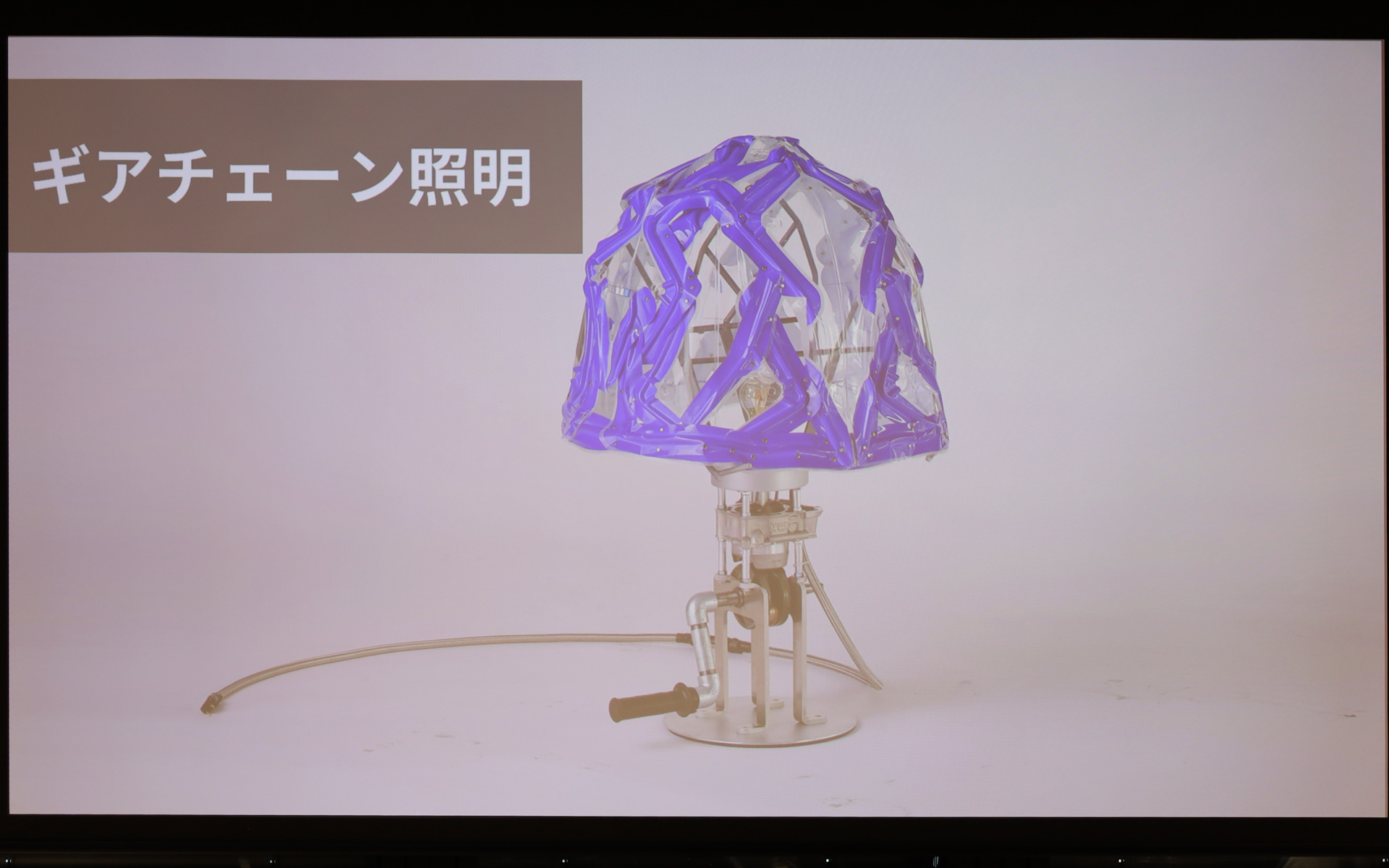 ただ照らすだけではない、ヤマハ発動機のエンジンの機能美に触れて学べる照明。8mm厚のアルミ一体成形で作られた土台には、本物の4ストロークエンジンの機構を搭載。ハンドルを手で回すとギヤが噛み合い、ピストンが往復。実際のエンジン同様、圧縮・爆発のタイミングに合わせてLEDが点灯するギミックを組み込んだ。光を拡散するシェード部分には、塗装工程で使われた「ハンガー（吊り具）」を骨組みにして、タンク用デカールの端材をパッチワークのように配置。廃材の隙間から漏れる不規則な光が、エンジンの鼓動をドラマチックに演出する