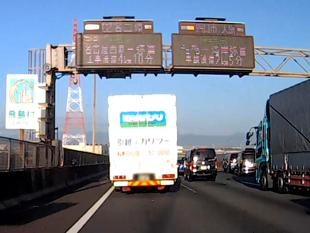 車線変更するトラックの先、追い越し車線で玉突き事故。真上の電光掲示板は「ここから－湾岸弥富 事故渋滞 2km 5分」の表示