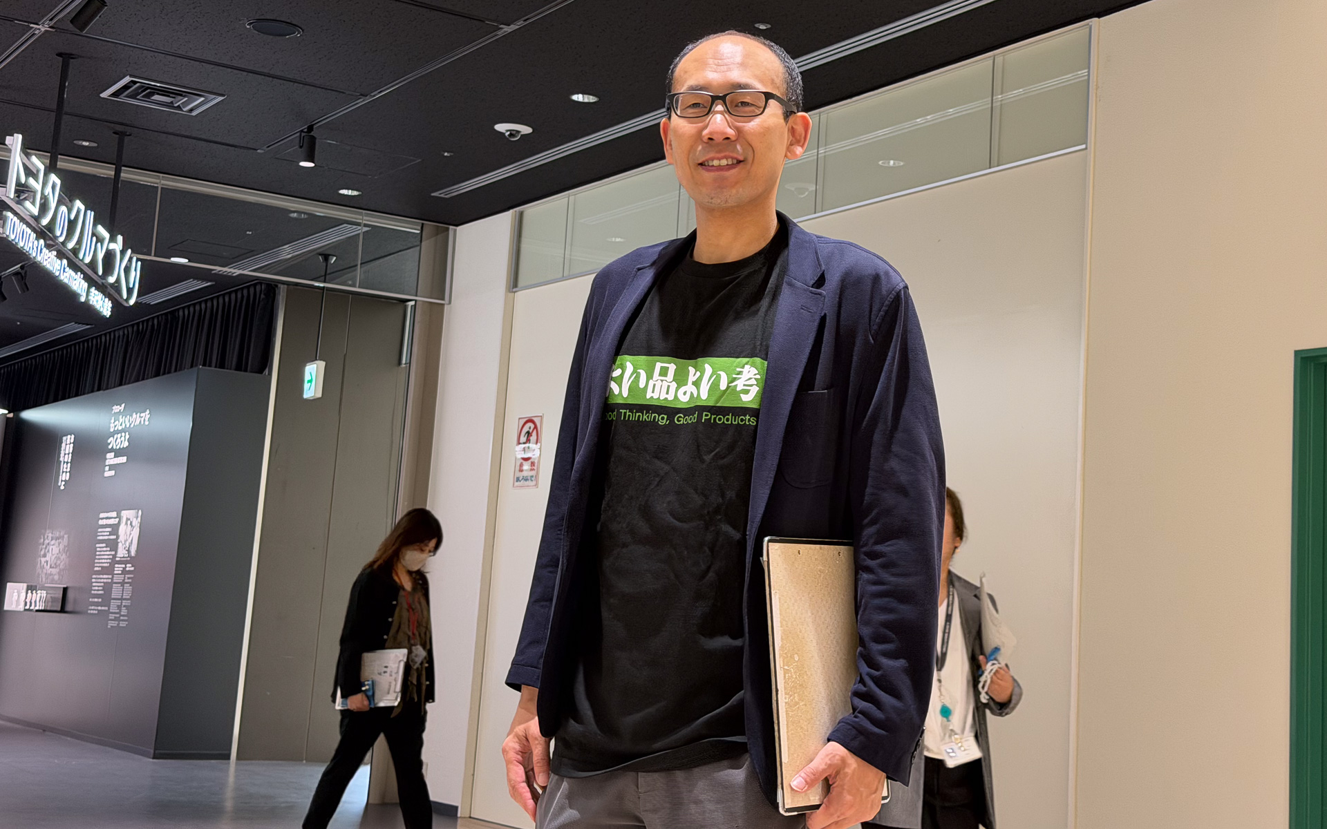 トヨタ会館の新しいおみやげ、「よい品よい考」Tシャツ。モデルは新展示「トヨタのクルマづくり」に携わったトヨタ自動車株式会社 社会貢献部 企業・車文化室 会館1グループ長 諸留逸氏。紺のジャケットと合わせての着こなし