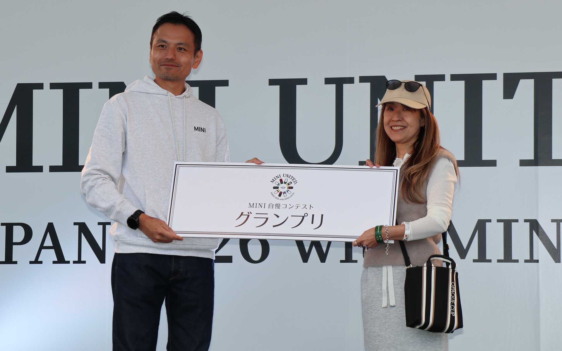 ビー・エム・ダブリュー株式会社 MINI本部長 向井健二氏（左）から記念のボードと「MINI PAUL SMITH BAG COLLECTION」トートバッグがMINTさん（右）に手渡された