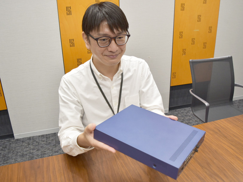 早坂智暁氏（コミュニケーション事業部 CC開発部 ハードウェアグループ 主事）。筐体が大型化しているのが見て取れるが、その分放熱に寄与したほか、スイッチと同サイズにしたことで、製造効率やコスト効率の向上などのメリットも得られたという