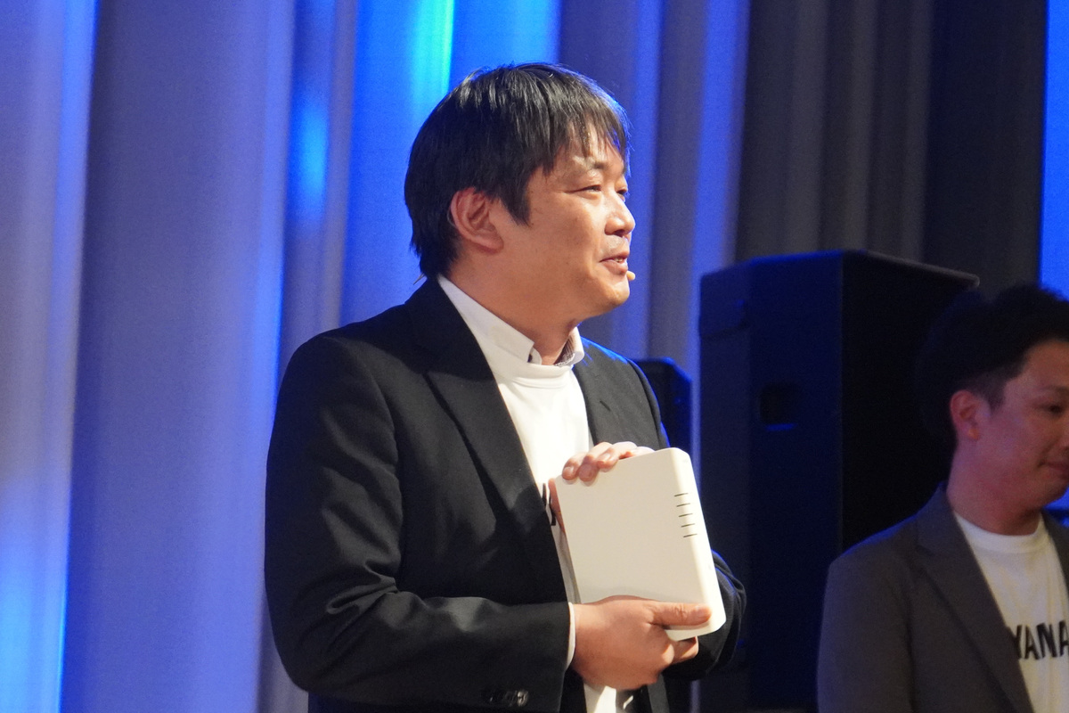 SCSK株式会社の山科正幸氏（ITインフラサービス事業グループ ネットワーク事業本部 ネットワークプロダクト第一部 技術課 課長代理）。NWR100を手に持っている