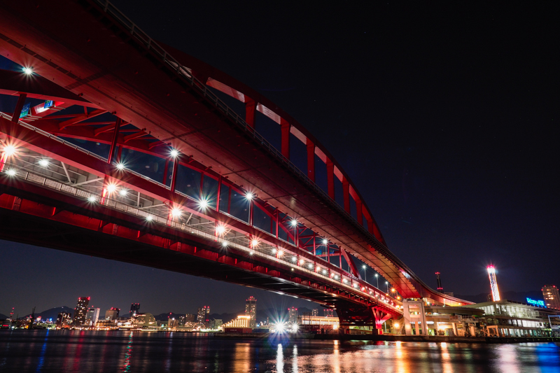 大橋の先に広がる港町。徒歩だと渡り切るのに20分ほどかかる橋だ。夜の光が海面に反射することで、さらに輝きを増していた。人工光の色が鮮やかに表現できた。<br><span class="fnt-85">オリンパス PEN-F / M.ZUIKO DIGITAL ED 7-14mm F2.8 PRO / 11mm（22mm相当） / マニュアル露出（F8、13秒） / ISO 400</span>