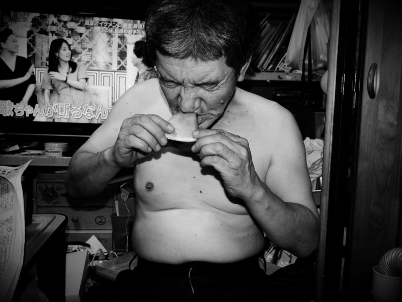 今夏初めてのスイカにかぶりつく父。家にはクーラーがないので、夏はいつも半裸ですごしている。ストロボを使って光量を調整した。<br><span class="fnt-85">オリンパス PEN-F / M.ZUIKO DIGITAL 17mm F1.8 / 17mm（34mm相当） /  マニュアル露出（F5、1/60秒） / ISO 800/ WB:オート</span>