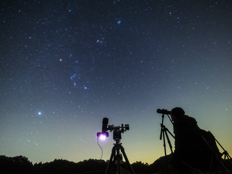 今回は視界の広いR1駐車場に赤道儀をセットして星雲や星団の撮影をした。画面中央の機材が望遠レンズでプレアデス星団を追尾撮影している赤道儀だ。右はイスに腰掛けてカメラ三脚に固定した口径70mm14倍の大型双眼鏡でオリオン大星雲を観望中の取材スタッフ。センターポールの傾けられる三脚だと双眼鏡が覗きやすくて楽チン。<br>OLYMPUS OM-D E-M5 Mark II／M.ZUIKO DIGITAL ED 7-14mm F2.8 PRO／ISO3200／7mm／F2.8開放／30秒／LEEソフトフィルターNo.3使用